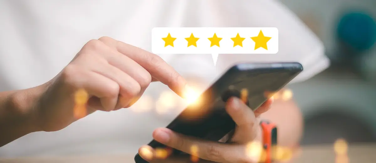 Nahaufnahme von zwei Händen, Eine Hand hält ein Smartphone und die andere bedient es mit dem Zeigefinger. In der Bildmitte ist ein weißer Balken oberhalb des Smartphones in dem fünf Sterne in Gold sind. Im Hintergrund ist ein weißes T-Shirt zu erkennen. Über das Bild sind kleine Leuchtelemente verteilt.