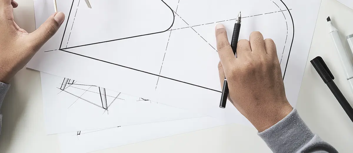 Nahaufnahme von Händen, die auf einem technischen Zeichnungspapier arbeiten. Eine Hand hält einen Stift und zeigt auf eine Linie, während die andere ein Zeichenwerkzeug hält Die Zeichnung besteht aus geometrischen Formen, Linien und Markierungen. Im Hintergrund sind weitere Zeichenwerkzeuge wie ein Filzstift und ein Marker zu sehen.