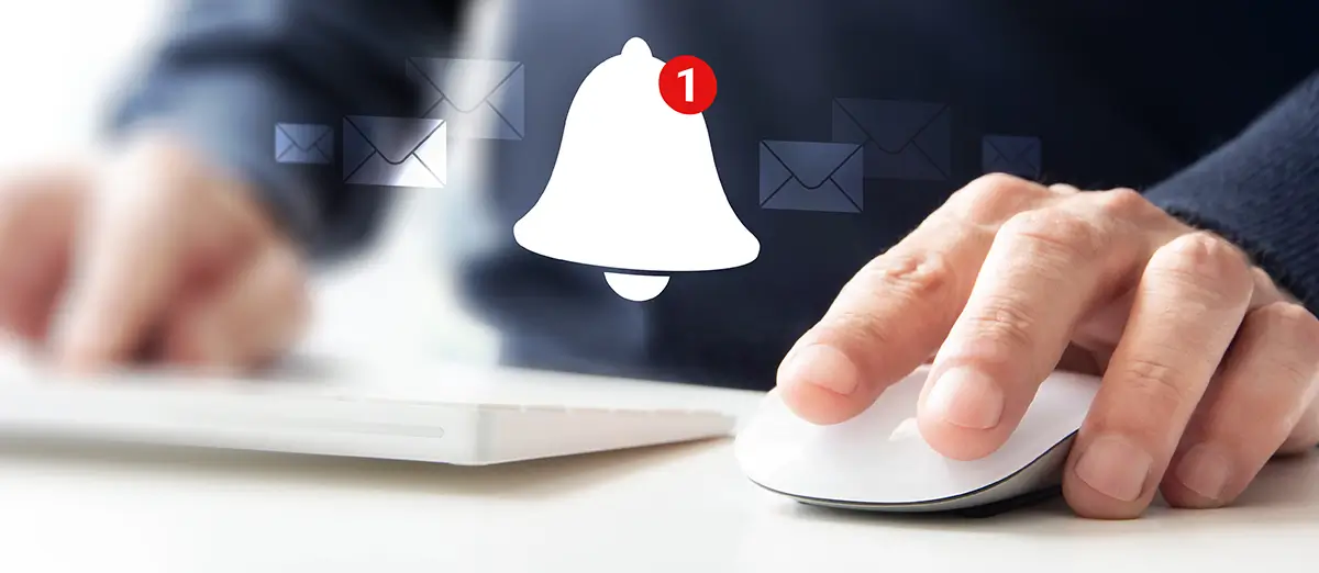 Nahaufnahme von zwei Händen, eine Computer-Tastatur und eine Computer-Maus am unteren Bildrand bedienen. Im Hintergrund ist eine Person bis zur Bauchmitte zu sehen mit einem blauen Pullover. In der Bildmitte sind eine weiße Glocke und weiße Briefumschläge, sie verblassen zu den Seiten.