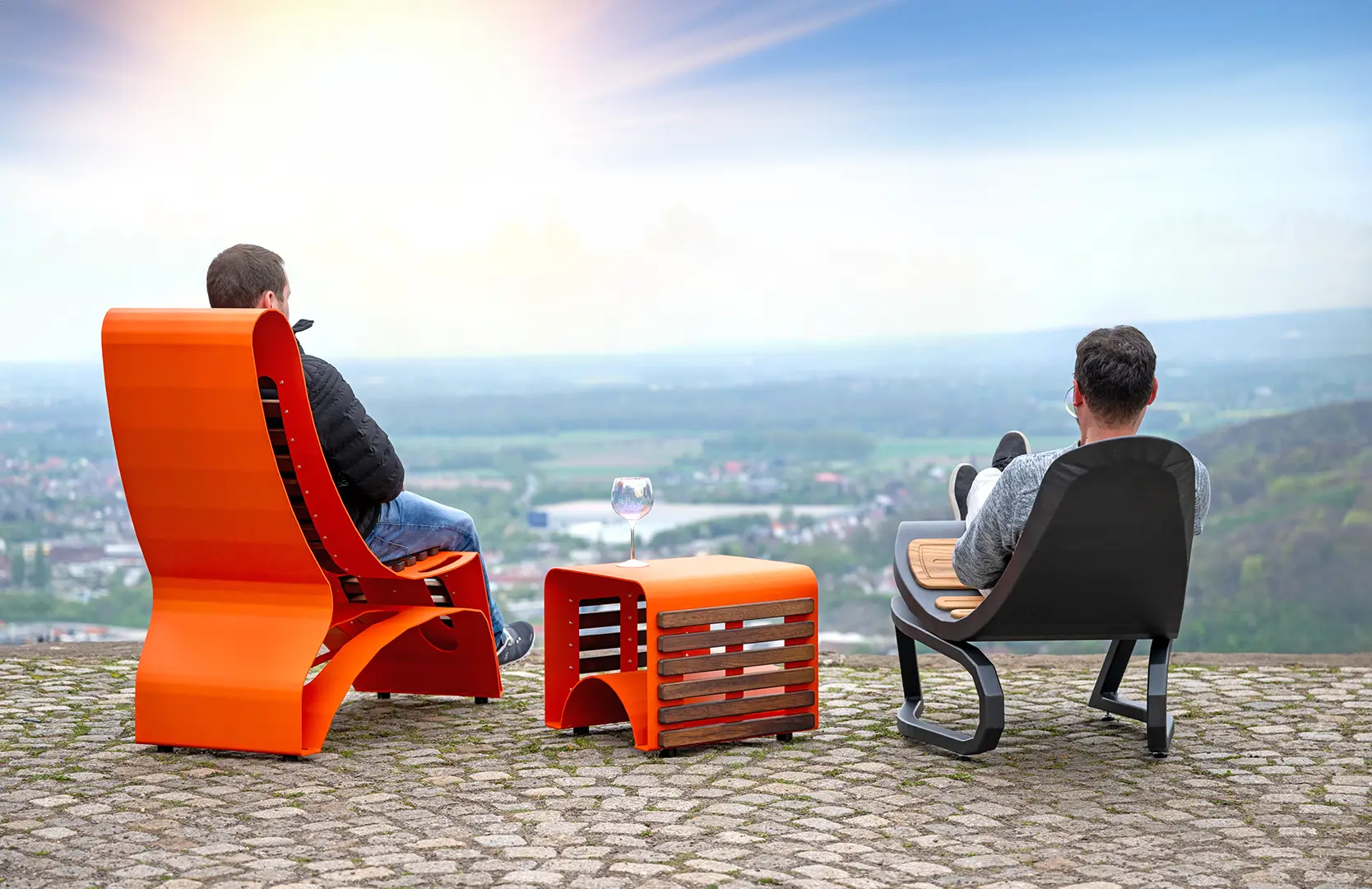Zwei Personen sitzen auf modernen, orangefarbenen und schwarzen Designer Liege Sesseln mit einem passenden Tisch in Orange in der Mitte, im Hintergrund eine weite Landschaft mit Natur und klarem Himmel
