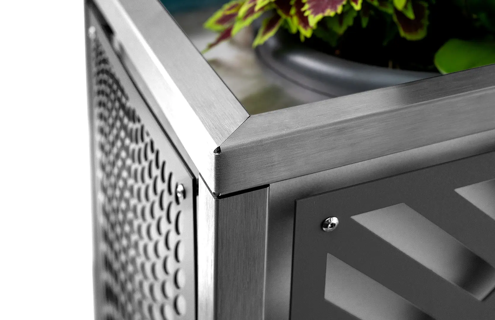 Detailaufnahme eines Pflanzkübels aus Metall in silber und schwarz, das schwarze Element ist mit zwei sichtbaren Schrauben mit dem Rahmen verschraubt und wird von fließenden Formen ähnlich wie Kieselsteine unterbrochen, in der Ecke des Bildes sieht man ein paar grün und rote Pflanzenblätter